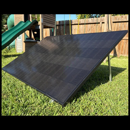 Black bifacial solar panel tilted on a ground-mount rack in a sunny backyard with a wooden fence and play set