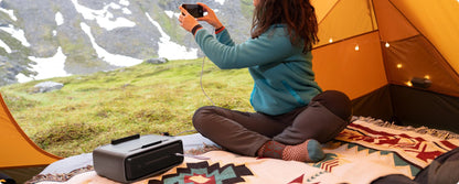 Person sitting inside a tent taking a photo of a scenic landscape while using a smart battery to charge the smartphone.