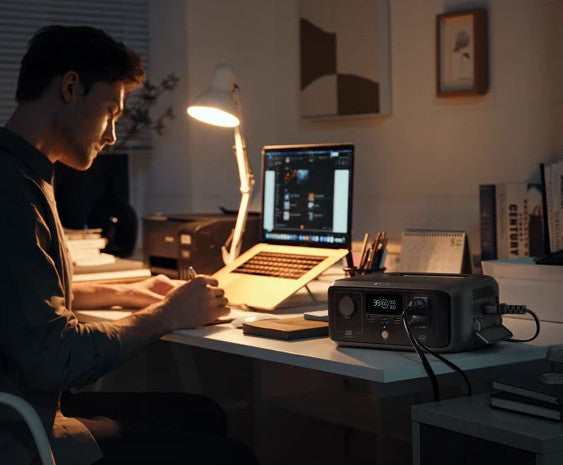 Person working at a desk with a laptop and portable power station in a dimly lit room.